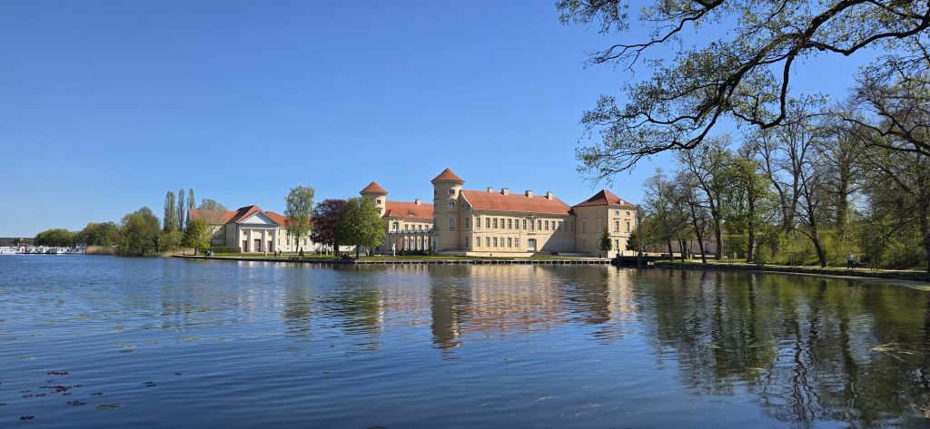 Der Tag beginnt direkt am beeindruckenden Schloss Rheinsberg, das idyllisch am Wasser liegt. Schon beim Ankommen merkt man, wie besonders dieser Ort ist.
Das Schloss bildet den perfekten Ausgangspunkt für einen Spaziergang durch die Umgebung.
Der Rheinsberg Tagesausflug zeigt besonders im Schlosspark seine Vielfalt., denn es ist der zentrale Ausgangspunkt des Ausflugs und besonders am Wochenende ein beliebtes Ziel. Gerade bei gutem Wetter kann es hier rund um den Schlossbereich etwas voller werden. Hier lohnt es sich in den frühen Morgenstunden zu starten.
Wer es etwas ruhiger mag, sollte einfach ein paar Schritte weiter in den Schlosspark gehen – dort verteilt sich der Besucherandrang schnell, und man findet deutlich mehr Ruhe inmitten der Natur.
Das Schloss selbst kann übrigens auch von innen besichtigt werden. Wir haben das an diesem Tag jedoch nicht gemacht und uns stattdessen für einen Spaziergang durch den Park entschieden.